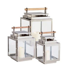 Three Adwin Outdoor Lanterns with silver frames, glass panels, and teak handles, each holding a white pillar candle, are displayed together in small, medium, and large sizes.