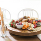 A Classic Trivet Top Lazy Susan, handcrafted from reclaimed wood, holds a variety of food items including fruits, pastries, cheese, and crackers, all arranged neatly. A bottle with breadsticks and empty white chairs are in the background.