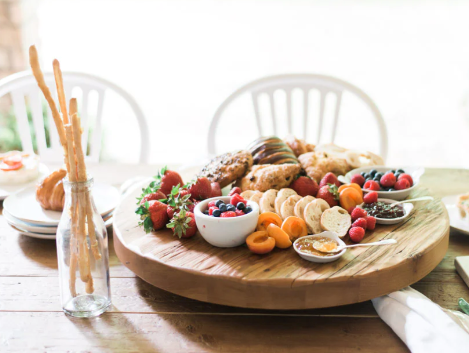 A Classic Trivet Top Lazy Susan, handcrafted from reclaimed wood, holds a variety of food items including fruits, pastries, cheese, and crackers, all arranged neatly. A bottle with breadsticks and empty white chairs are in the background.