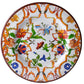 The Mario Luca Giusti Pancale White Collection showcases a decorative ceramic plate adorned with a vibrant floral pattern in shades of orange, green, blue, and red on a white background.