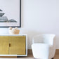 A Samantha Swivel Chair sits next to a gold-colored cabinet topped with a decorative item, below a framed abstract artwork on a white wall.