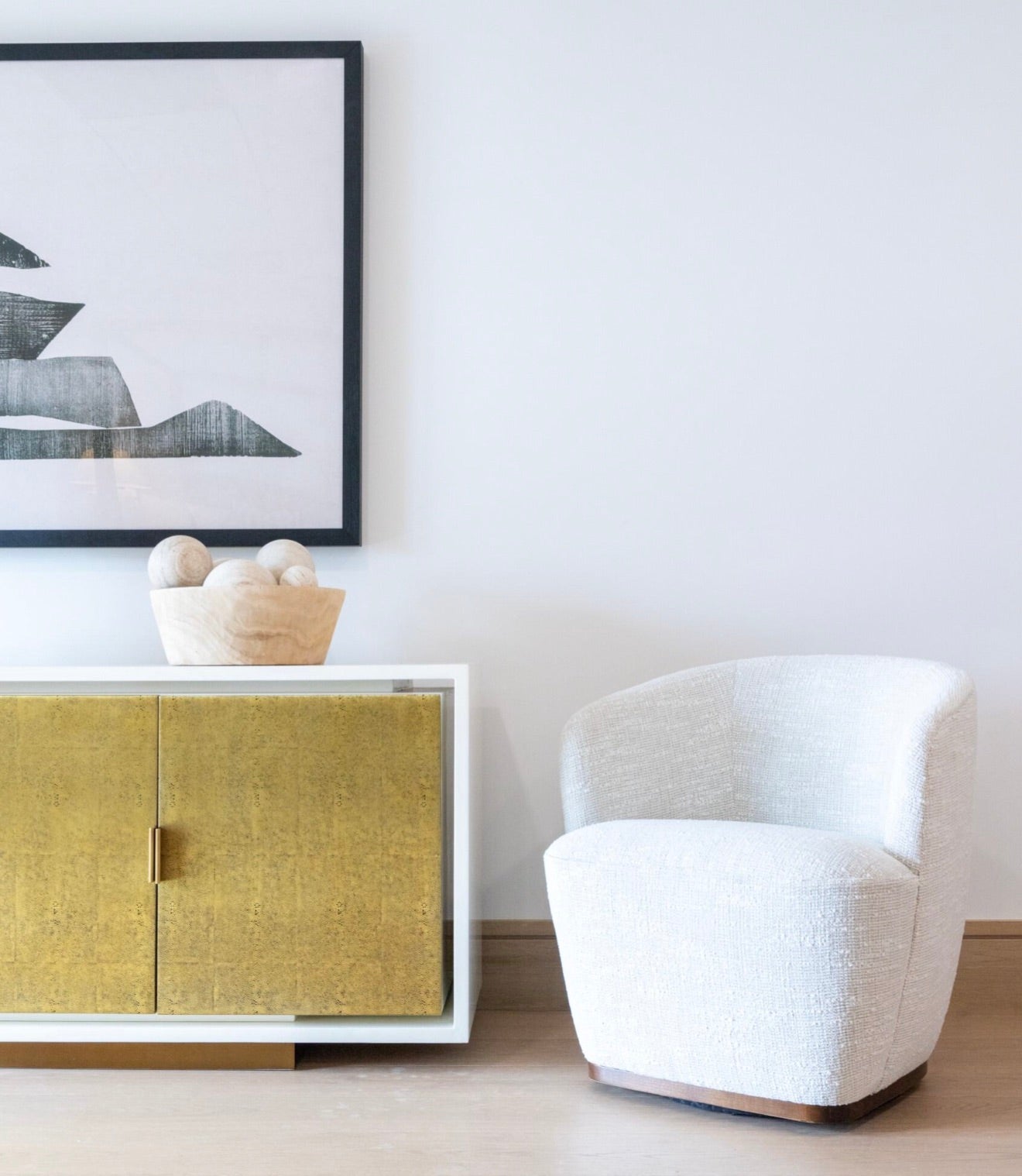 A Samantha Swivel Chair sits next to a gold-colored cabinet topped with a decorative item, below a framed abstract artwork on a white wall.