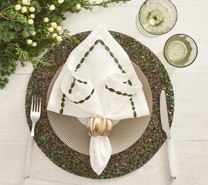 A festive place setting features a white plate, green beaded placemat, silverware, two green glasses, and greenery on a white table with an elegant Kim Seybert Swag Napkin Set in a gold ring.