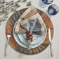 A table setting with a speckled plate, patterned charger, Kim Seybert Brown & Gold Tendril Napkin Ring holding a folded napkin, fork, knife, two glasses, and a floral branch on light wood—elegance meets nature.