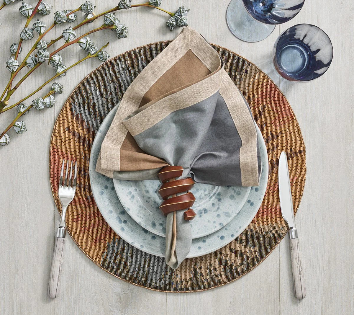 A table setting with a speckled plate, patterned charger, Kim Seybert Brown & Gold Tendril Napkin Ring holding a folded napkin, fork, knife, two glasses, and a floral branch on light wood—elegance meets nature.