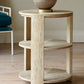 A round Tribeca Side Table, Bleached Aged Oak with curved legs and two shelves, featuring a beige decorative vase on top, stands proudly on a light-colored wooden floor next to a teal upholstered chair.