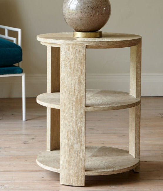 A round Tribeca Side Table, Bleached Aged Oak with curved legs and two shelves, featuring a beige decorative vase on top, stands proudly on a light-colored wooden floor next to a teal upholstered chair.