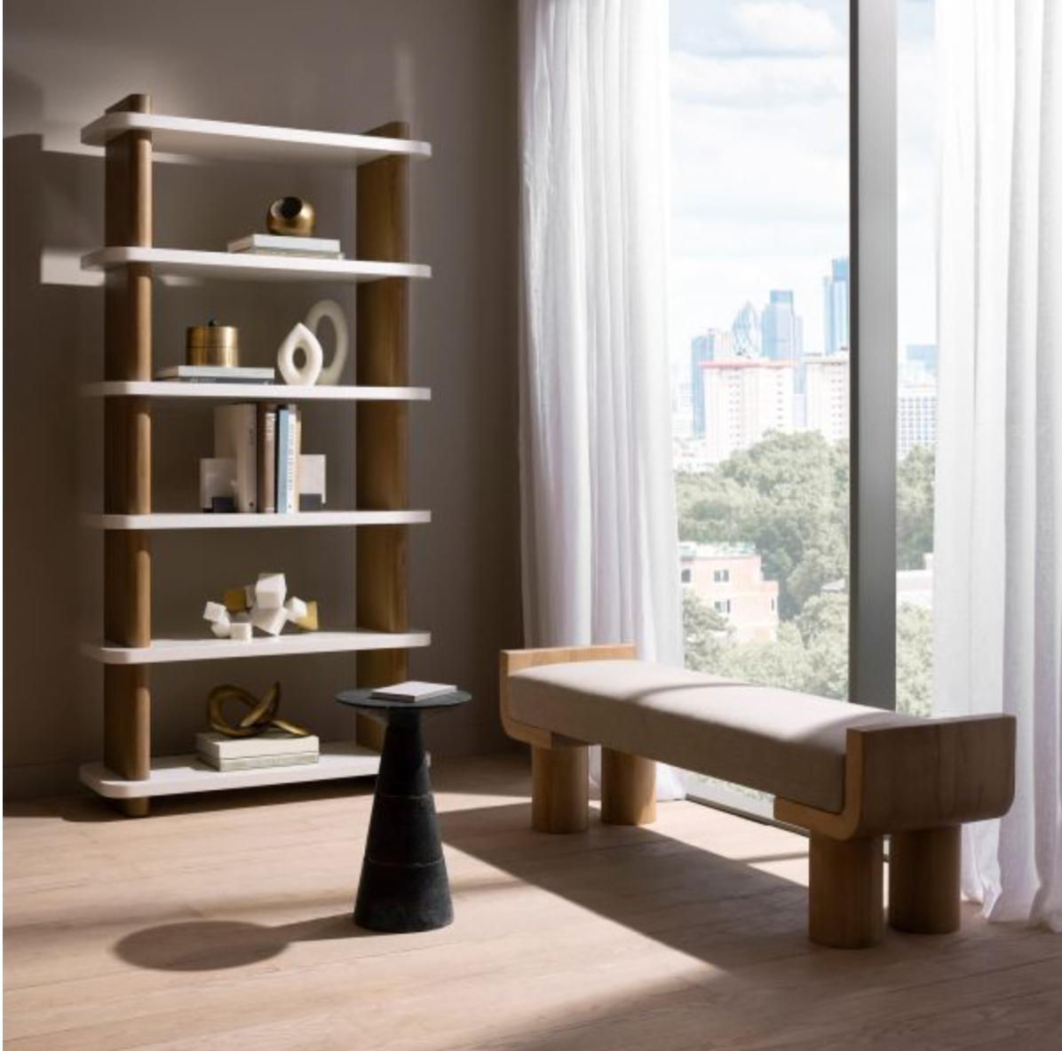 A modern living space with a tall Triton Bookshelf, Oyster featuring matte white lacquer shelves, a small geometric table, and a cushioned bench. The room has light wooden floors and large windows with white curtains, showcasing a stunning city skyline view.