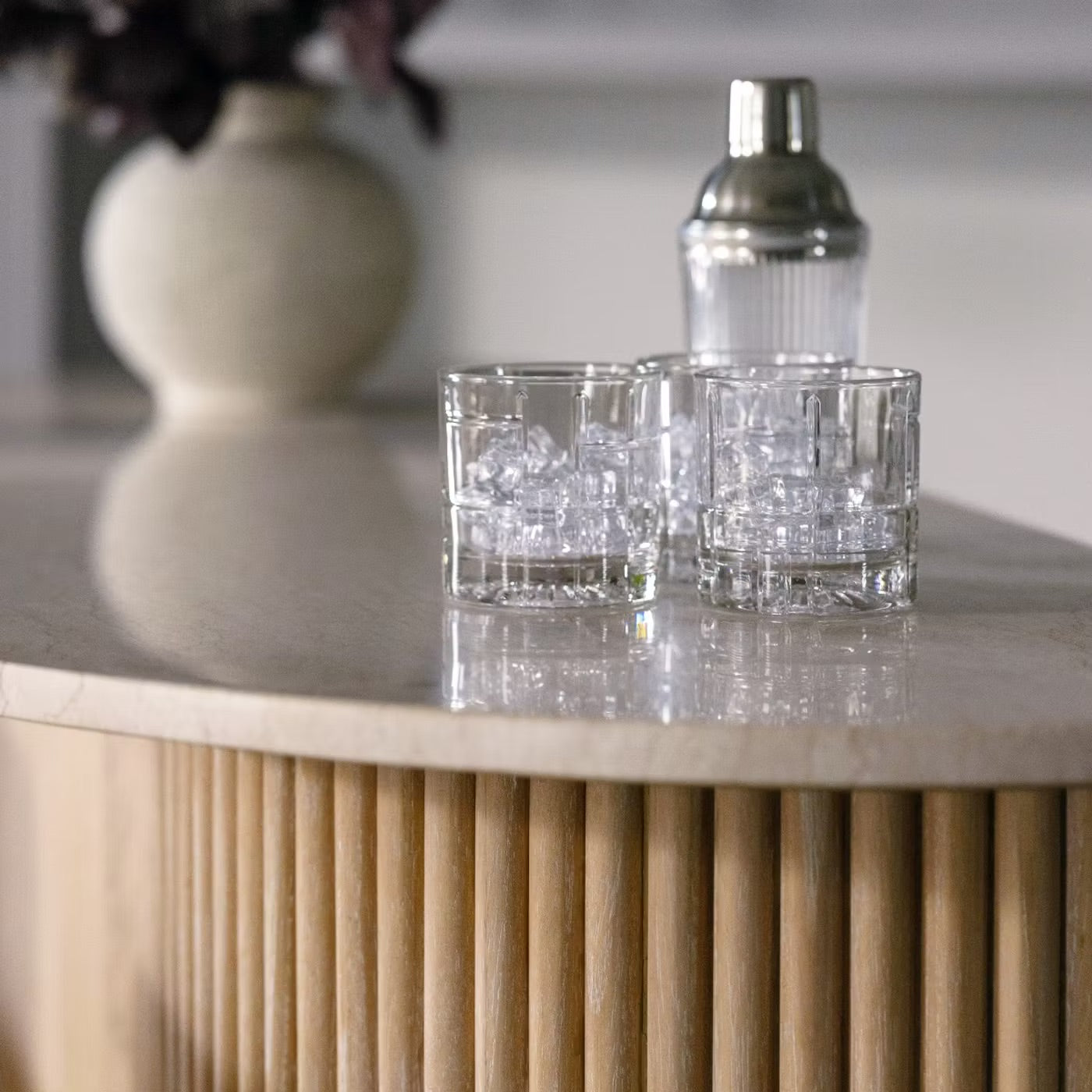 Two glasses with ice cubes and a cocktail shaker rest on the light wood Anton Bar, which features clever storage solutions and is accented by a vase in the background.