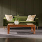 The Bradford Coffee Table, featuring a brown patterned finish and topped with a vase of green foliage, sits on a gray rug in front of a green velvet sofa with two cream pillows—bringing elegance to any room with dark paneled walls.