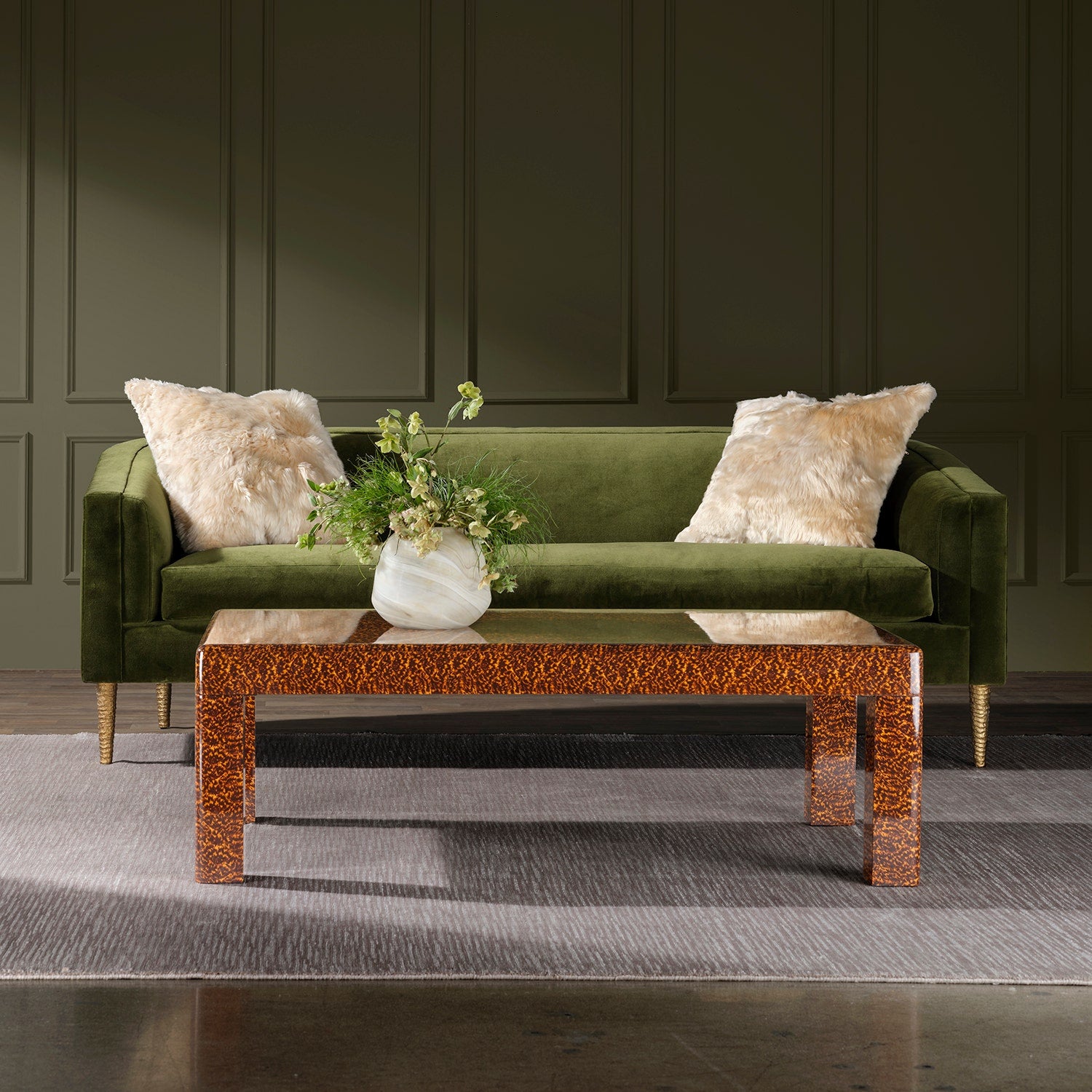 The Bradford Coffee Table, featuring a brown patterned finish and topped with a vase of green foliage, sits on a gray rug in front of a green velvet sofa with two cream pillows—bringing elegance to any room with dark paneled walls.
