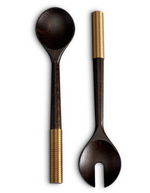 Two hand-carved dark wooden salad servers with gold-wrapped handles, one featuring a round spoon and the other a forked tip, placed side by side on a white background. These elegant serving utensils are crafted from smoked ash for a touch of sophistication. The L'Objet Alhambra Serving Set adds an air of refined elegance to your dining experience.