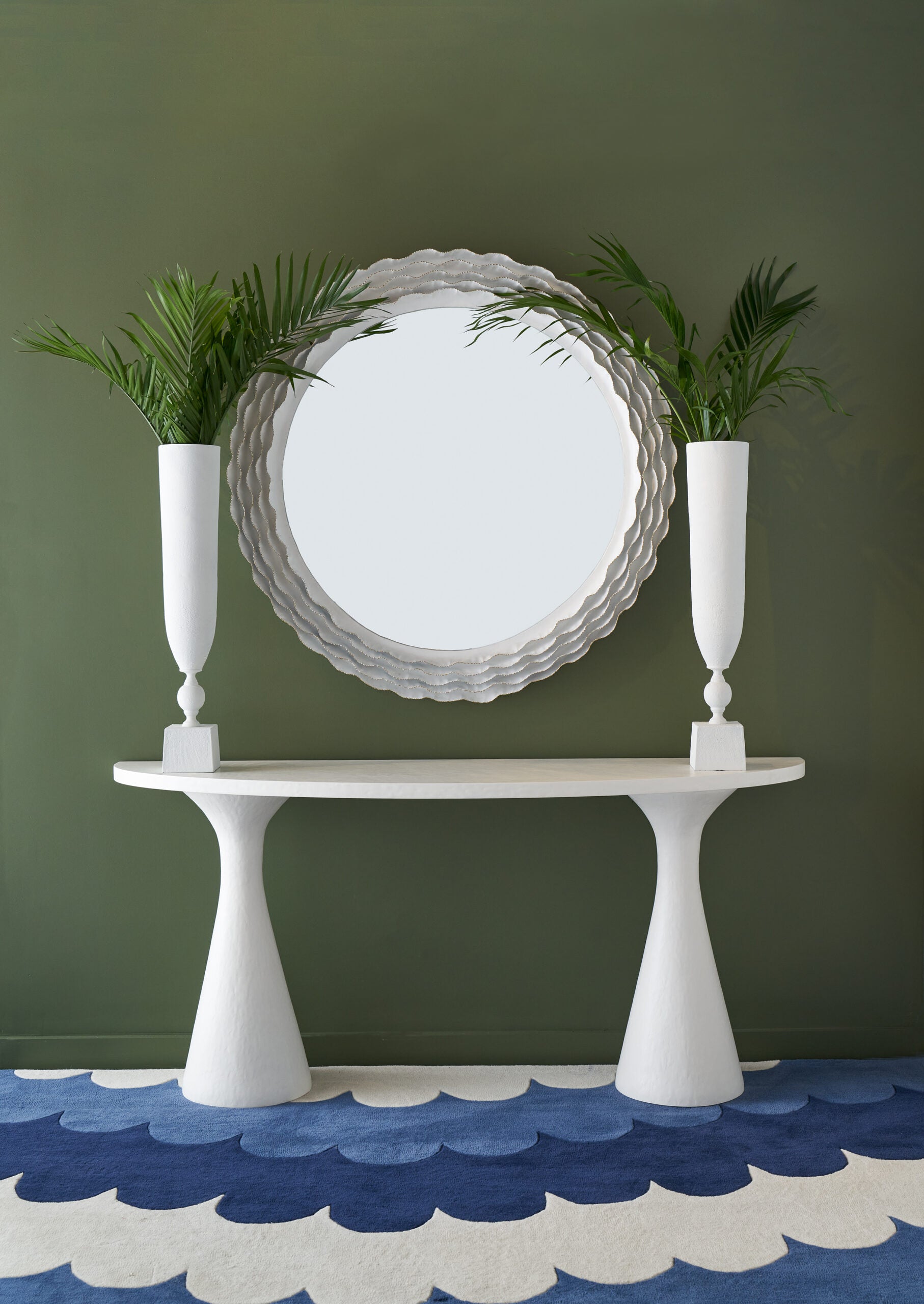 A white Kasper Demilune Console Table sits against an olive green wall, topped with two tall vases of palm leaves, while a round scalloped mirror and a blue wave-patterned rug complete the stylish entryway.
