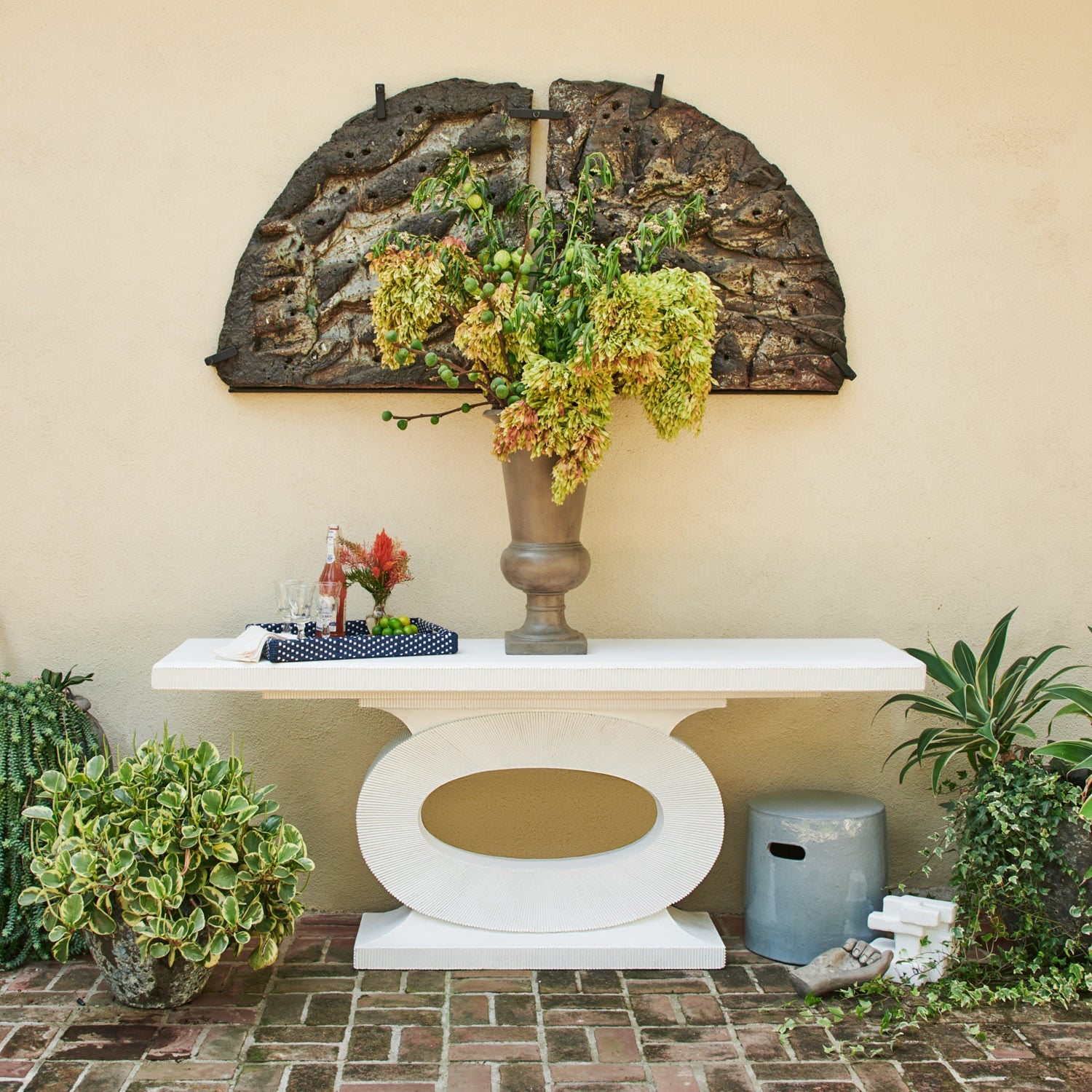 The Grier Console, featuring a white sculptural design with an oval cutout, stands against a beige wall. It's topped with a large vase of green hydrangeas and surrounded by rustic wall art and potted plants.