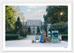 A young girl stands beside a Nick Mele Fine Art Champagne Stand and a collection of limited edition clothing and items for sale, set up in front of a large, gated mansion surrounded by trees in Newport, RI.